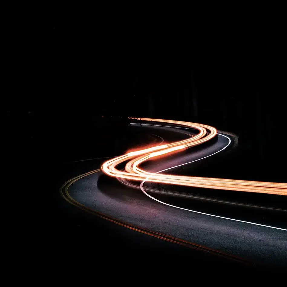 Long exposure photograph of a winding road at night with vehicle light trails creating smooth, flowing lines.
