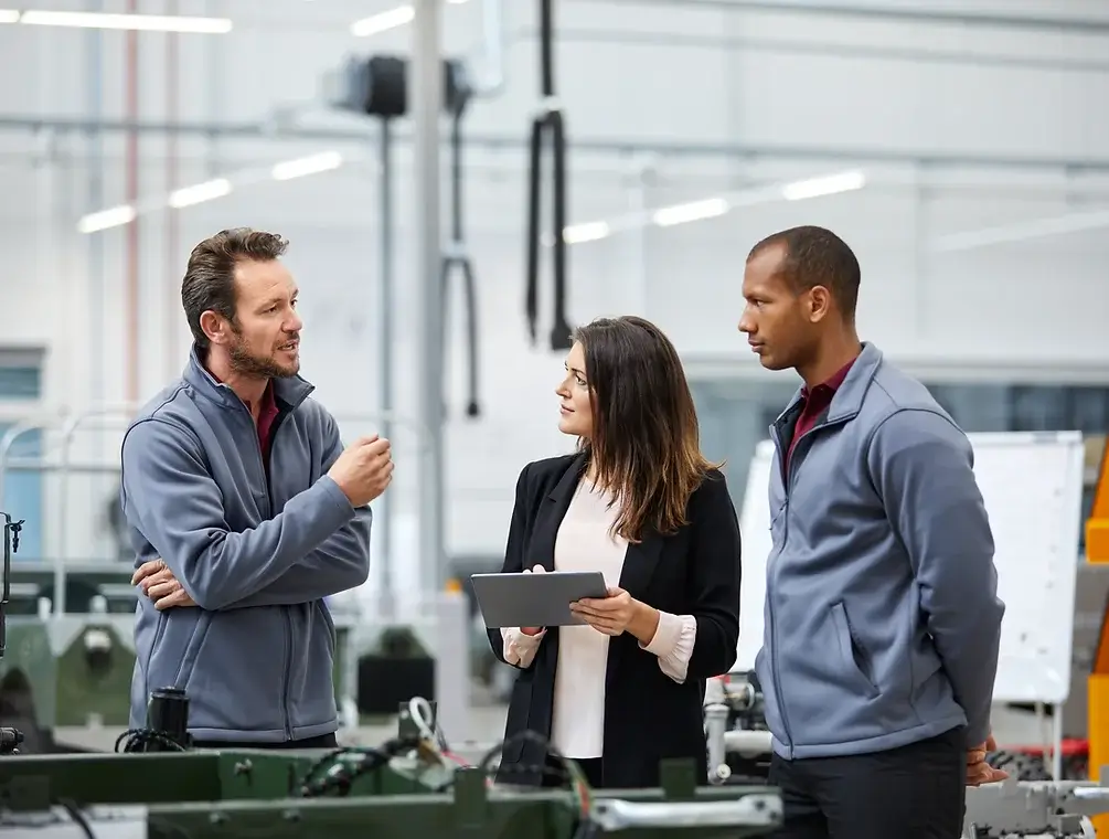 Three individuals in an industrial setting: a man on the left speaking and gesturing, a girl in the middle holding a tablet, and a man on the right wearing a red and grey jacket.